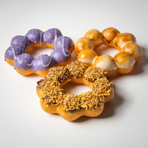 Three mochi donuts: purple with sprinkles, glazed, and chocolate with toasted coconut flakes.