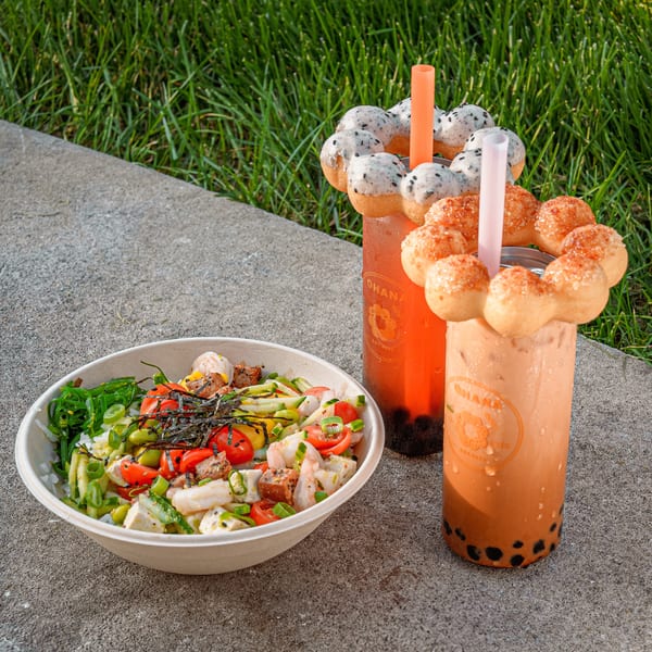 A poke bowl with two boba drinks topped with mochi donuts on a concrete path next to grass.
