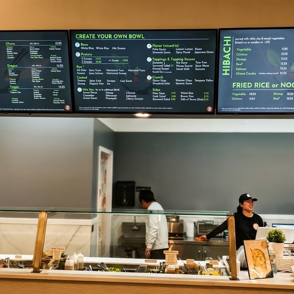 Digital menu boards display food options above a restaurant counter with staff and fresh ingredients.