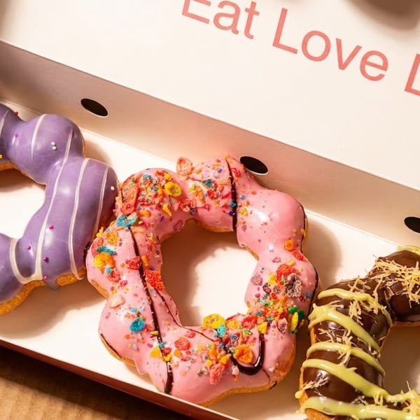 Three colorful mochi donuts: purple, pink with cereal, and chocolate, in a box with 'Eat Love Life' text.