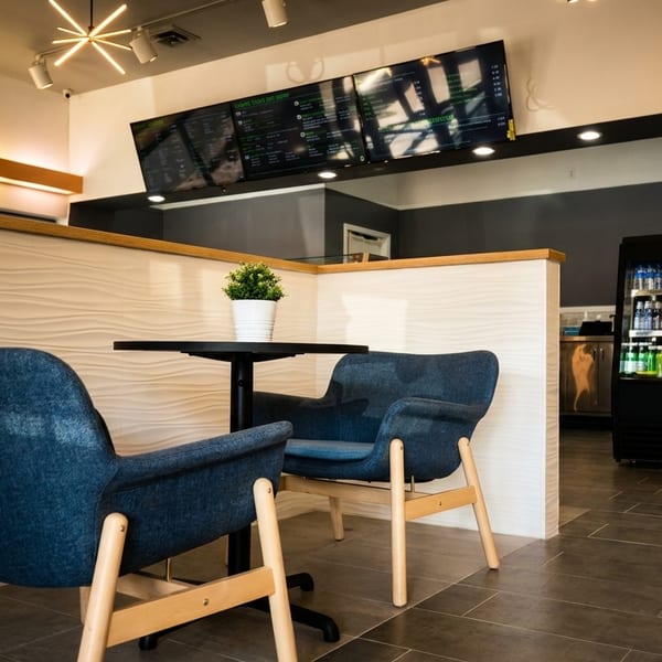 Cozy restaurant interior with two blue armchairs, a small table, menu boards, and a drink cooler.