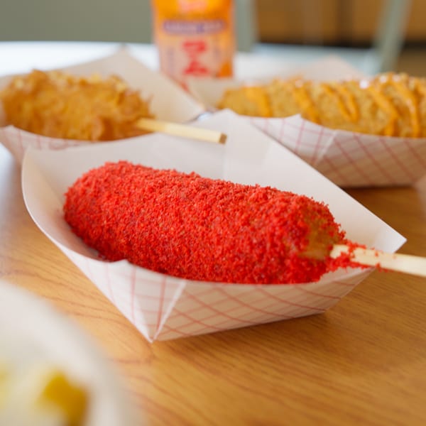 A bright red, Flamin' Hot Cheetos-dusted corn dog in a paper tray, with other corn dogs blurred in the background.