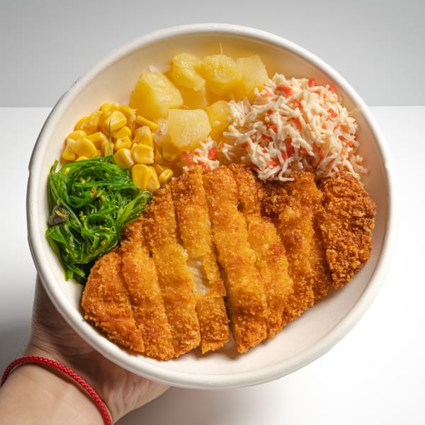 Hand holding a bowl with sliced fried chicken, seaweed salad, corn, pineapple, and coleslaw.